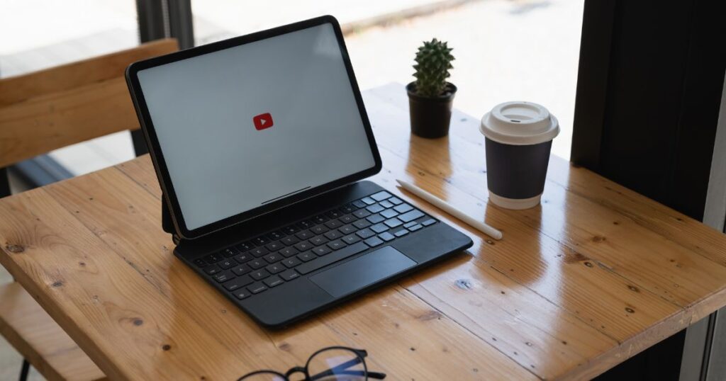 A tablet or iPad with keyboard on a small wooden table. The small red YouTube icon is on the screen.