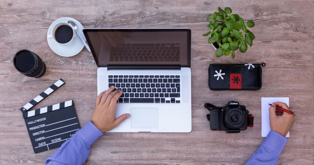Overhead view of a person at a table working on a laptop. A digital camera is to the right of the laptop and a small clapperboard is to the left of the laptop.