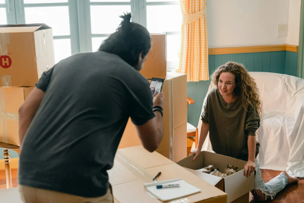 A woman posing for a photo a man is taking while she is packing a box.