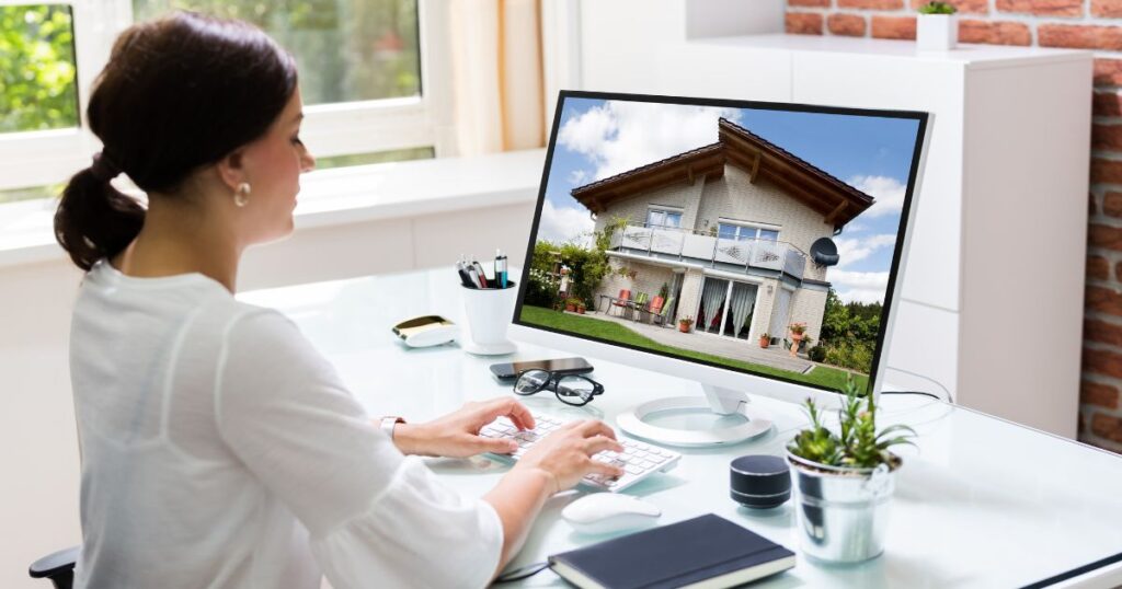 A woman at a desk typing on a keyboard in front of a monitor displaying an image of a house.