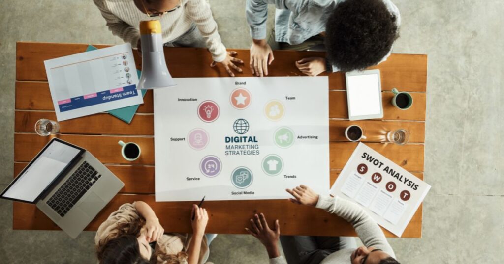 Overhead view of a team seated at a table for a digital marketing meeting, with marketing materials, a tablet, and a laptop on the table.