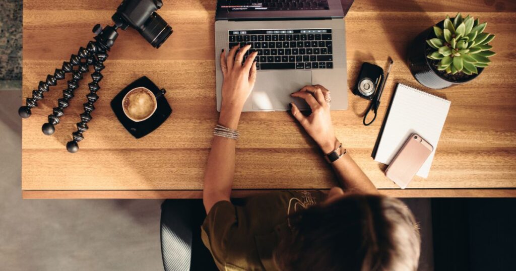 Overhead view of a woman at a desk typing on a laptop with a camera lying next to the laptop and a smartphone to the right of the laptop.