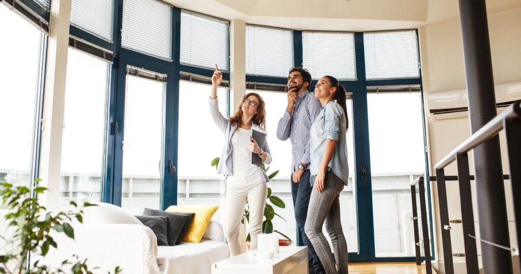 A real estate agent is pointing out a feature in a home to a smiling young couple.