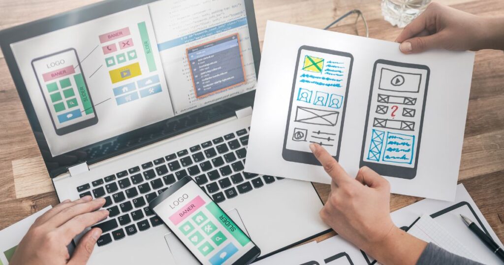 Overhead view of woman's hands holding and pointing to a paper showing smartphone app screen mock-up. Another person's hand is shown resting on a laptop keyboard.