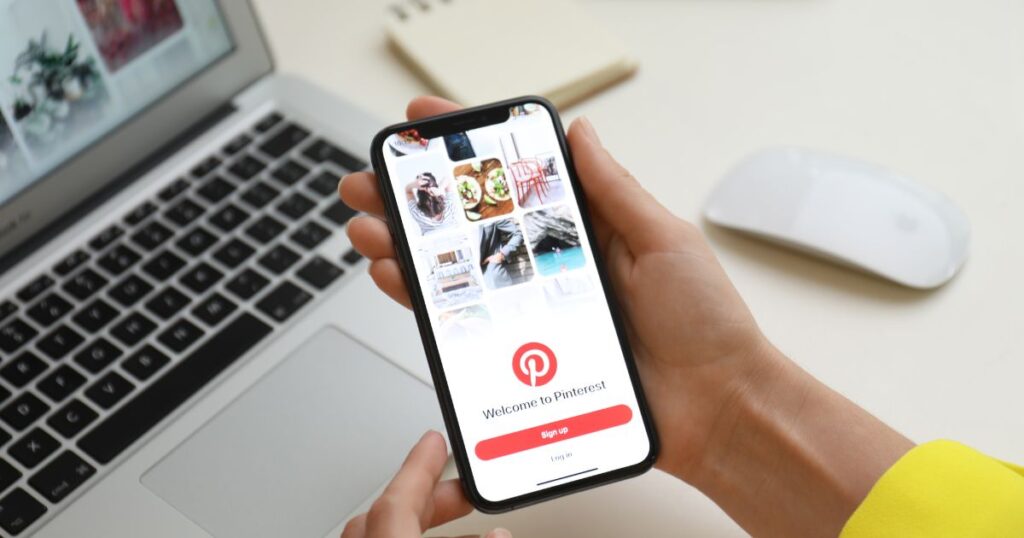 Closeup of a person's hands holding a smartphone with a Welcome to Pinterest page displayed on the phone's screen. There is a laptop and mouse in the background.