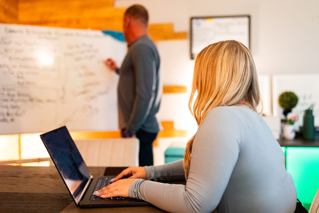 Woman with a laptop watching a man at a whiteboard in the background.