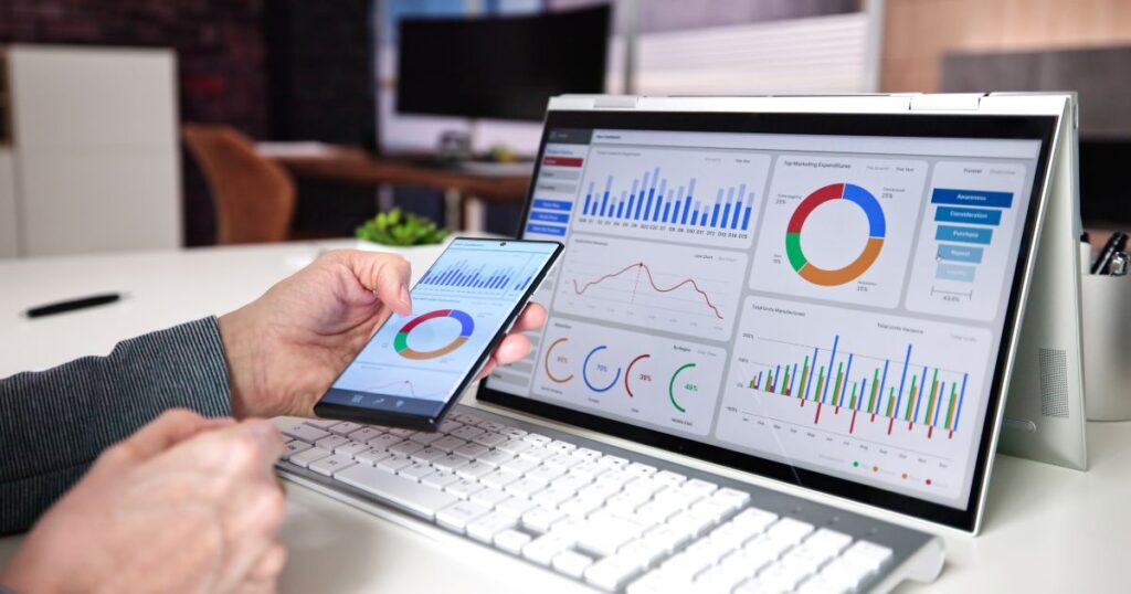Closeup of a person's hands holding a smartphone showing reporting analytics charts on the screen, with a table or iPad showing more detailed analytics charts and graphs on the screen.