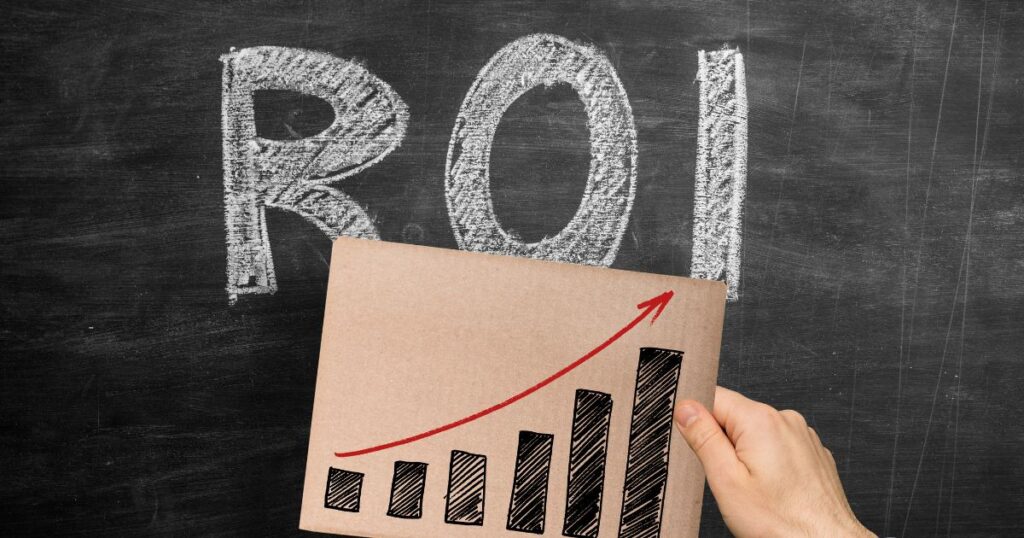 Closeup of a person's hand holding a cardboard square with a simple bar chart drawn on it to show an increase. There is a chalkboard with ROI drawn on it behind the cardboard square.