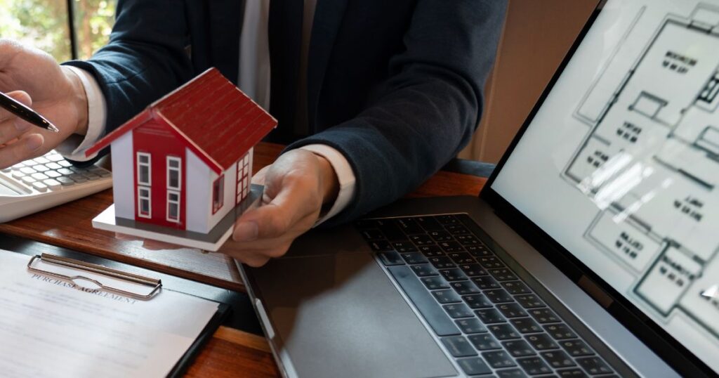 Unidentified man holding a model house with laptop nearby showing a floorplan on the screen.