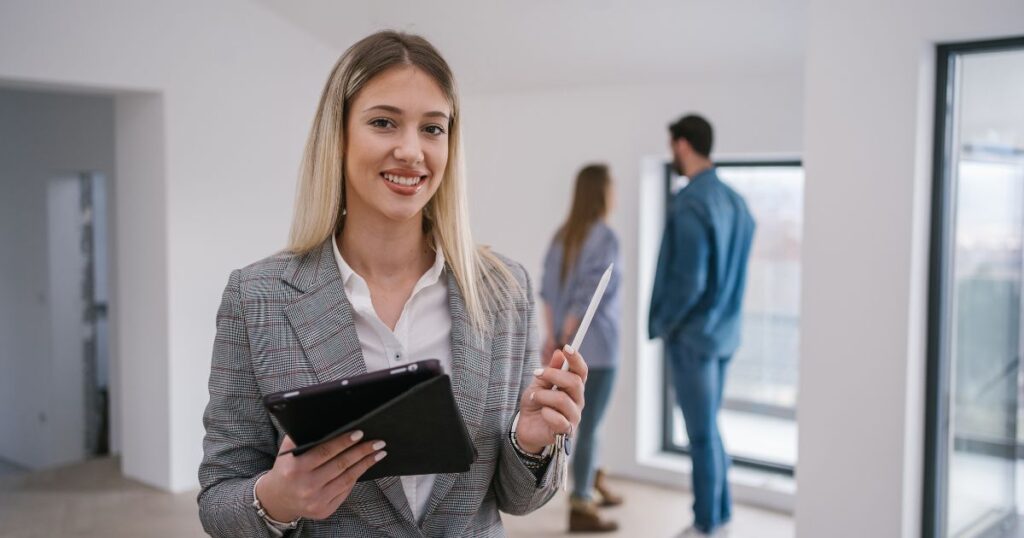 A smiling female real estate agent looking at the camera with clients across the room in the background. Knowing how to caption a real estate post on Instagram is key for social media marketing.